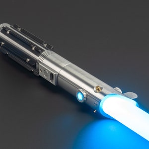 May include: A silver lightsaber with a blue glowing blade. The hilt is metallic with black accents and detailed mechanical components. The blade emits a bright, vibrant blue light, suggesting energy.