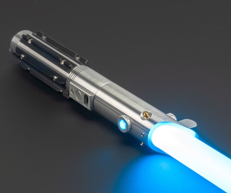May include: A silver lightsaber with a blue glowing blade. The hilt is metallic with black accents and detailed mechanical components. The blade emits a bright, vibrant blue light, suggesting energy.