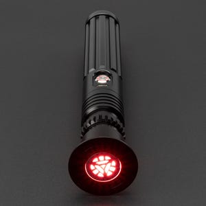 Op de afbeelding: Een zwart lightsaber met een rode, gloeiende, driehoekige emitter. De lightsaber heeft een getextureerd, cilindrisch lichaam en een zwarte, ronde handgreep.