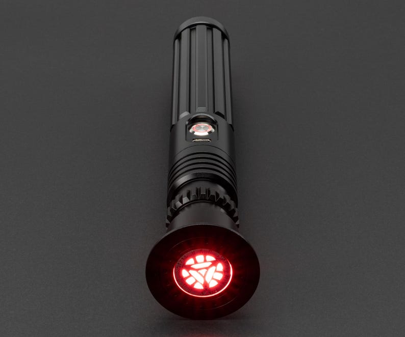 Op de afbeelding: Een zwart lightsaber met een rode, gloeiende, driehoekige emitter. De lightsaber heeft een getextureerd, cilindrisch lichaam en een zwarte, ronde handgreep.