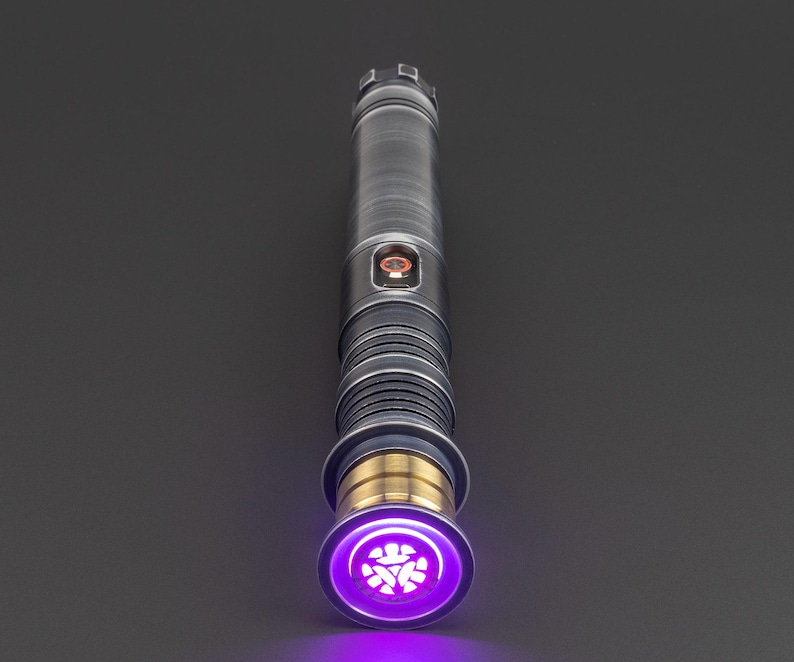 Puede incluir: Un sable de luz plateado con una hoja morada y un acento dorado. El sable de luz tiene un emisor circular con un s&iacute;mbolo en el centro.