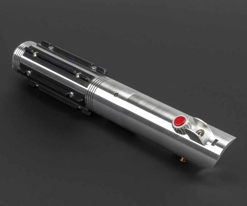 May include: A silver lightsaber with black accents and a red button. The hilt features a ribbed design and a curved end. The lightsaber is a prop or collectible item.