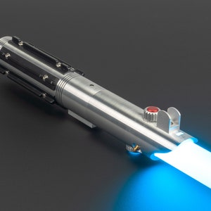 May include: A silver lightsaber with black accents and a red button. The lightsaber emits a bright blue beam of light, suggesting it is activated. The design is detailed and metallic, with a futuristic aesthetic.