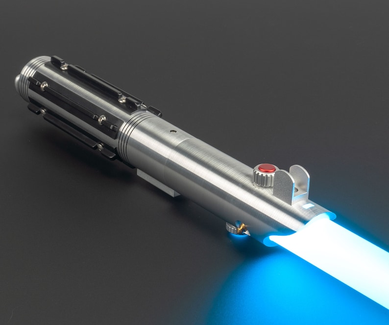 May include: A silver lightsaber with black accents and a red button. The lightsaber emits a bright blue beam of light, suggesting it is activated. The design is detailed and metallic, with a futuristic aesthetic.