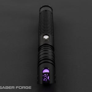 May include: A black lightsaber with a ribbed handle and a glowing purple emitter. The lightsaber has a silver button and the Saber Forge logo is visible at the bottom left.