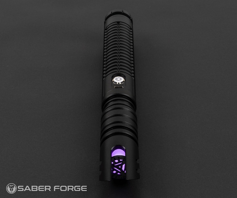 May include: A black lightsaber with a ribbed handle and a glowing purple emitter. The lightsaber has a silver button and the Saber Forge logo is visible at the bottom left.