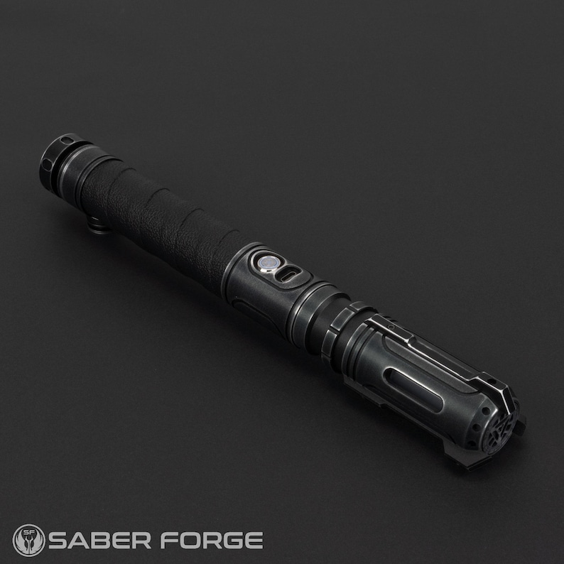 Pode incluir: Um sabre de luz preto com detalhes prateados. O punho tem uma pega texturizada e uma s&eacute;rie de bot&otilde;es e detalhes. A extremidade do sabre de luz tem um design detalhado. O logotipo Saber Forge &eacute; vis&iacute;vel.