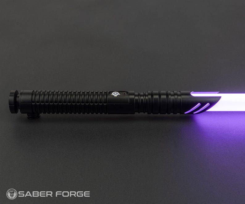 May include: A black lightsaber with a ribbed handle and a glowing purple blade. The hilt features a button and a logo. The lightsaber is set against a dark gray background. The Saber Forge logo is at the bottom.