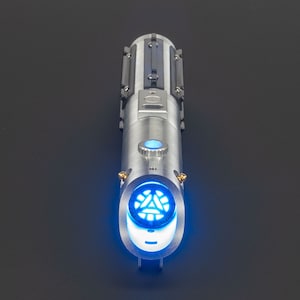 May include: A silver lightsaber with a blue glowing blade emitter. The hilt features a detailed design with black accents and a blue activation button. The end of the hilt has a circular design with a blue glowing pattern.
