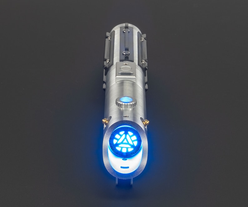 May include: A silver lightsaber with a blue glowing blade emitter. The hilt features a detailed design with black accents and a blue activation button. The end of the hilt has a circular design with a blue glowing pattern.