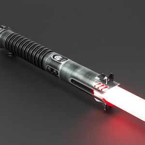 Op de afbeelding: Een zwart en zilveren lightsaber met een rode blade. De lightsaber heeft een getextureerde metalen afwerking en een kleine knop aan de zijkant.