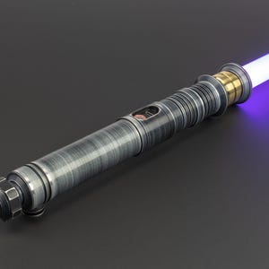 Puede incluir: Un sable de luz plateado con una hoja morada. El sable de luz tiene un mango negro con un acento dorado cerca de la hoja.