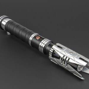 May include: A black and silver lightsaber with a textured grip and a detailed emitter. The lightsaber has a silver metal hilt with a black rubber grip.