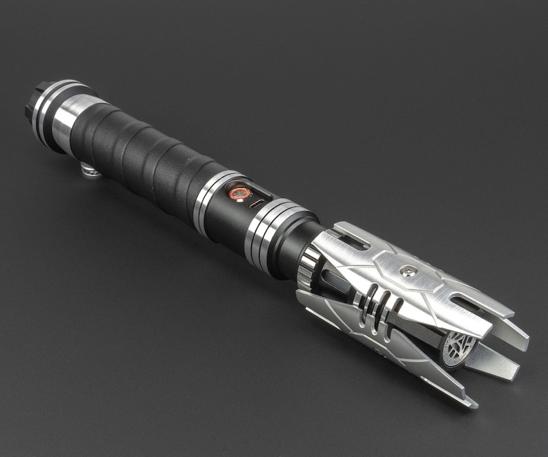 May include: A black and silver lightsaber with a textured grip and a detailed emitter. The lightsaber has a silver metal hilt with a black rubber grip.