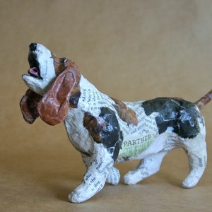 May include: A paper mache sculpture of a basset hound dog with brown, black, and white paper covering its body. The dog is standing on all fours with its head tilted back and mouth open. The paper is printed with text, including the words "Partner" and "Companion".