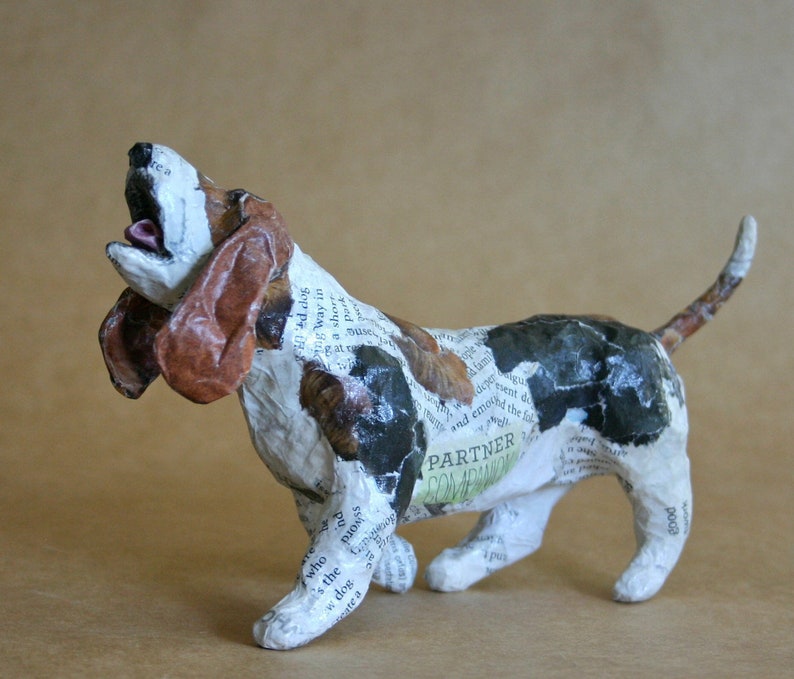 May include: A paper mache sculpture of a basset hound dog with brown, black, and white paper covering its body. The dog is standing on all fours with its head tilted back and mouth open. The paper is printed with text, including the words "Partner" and "Companion".