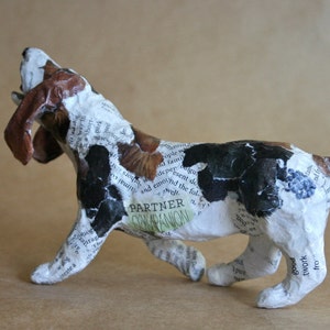 May include: A white, brown, and black paper mache sculpture of a dog with its mouth open, as if it is barking. The sculpture is covered in newspaper clippings and the words "Partner Companion" are visible on the dog's side.