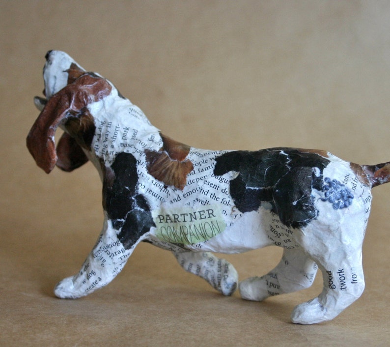May include: A white, brown, and black paper mache sculpture of a dog with its mouth open, as if it is barking. The sculpture is covered in newspaper clippings and the words "Partner Companion" are visible on the dog's side.