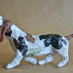 May include: A paper mache sculpture of a basset hound dog with a white, brown, and black patterned design. The dog is in a running pose with its mouth open and its tongue sticking out. The sculpture is covered in newspaper clippings and has the words "Partner Companion" written on its side.