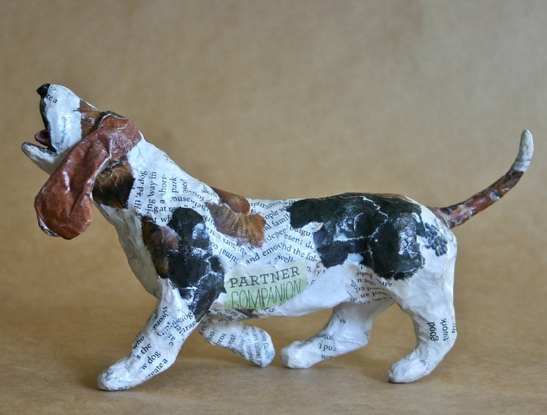 May include: A paper mache sculpture of a basset hound dog with a white, brown, and black patterned design. The dog is in a running pose with its mouth open and its tongue sticking out. The sculpture is covered in newspaper clippings and has the words "Partner Companion" written on its side.