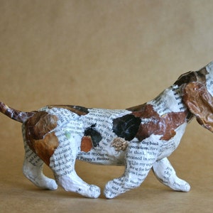May include: A paper mache sculpture of a brown and white basset hound dog with its mouth open, running. The sculpture is made of paper and is covered in printed text.