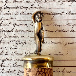 Może przedstawiać: Winiarski korek do butelki z mosiężną figurką małpy. Korek wykonany jest z korka i mosiądzu. Małpa jest w zabawnej pozie, z jedną ręką w górze. Tło to antyczny dokument.