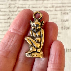 May include: A small, bronze-colored charm depicting a seated fairy or elf with pointed ears. The figure is detailed with visible facial features and is attached to a small loop at the top for hanging. The charm is likely made of metal.
