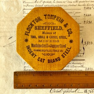 May include: An octagonal yellow wooden plaque with black text reading FLOCKTON, TOMPKIN & CO., SHEFFIELD. Makers of TOOL, DRILL & CHISEL STEEL, MINERS, Machine Drill & Jumper Steel, HAMMERS, FILES, TOOLS & co., INDENT CAT BRAND STEEL. A ruler is visible.