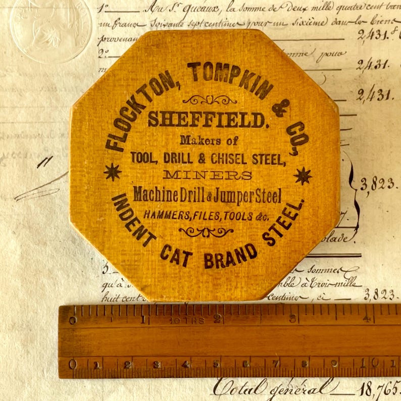 May include: An octagonal yellow wooden plaque with black text reading FLOCKTON, TOMPKIN & CO., SHEFFIELD. Makers of TOOL, DRILL & CHISEL STEEL, MINERS, Machine Drill & Jumper Steel, HAMMERS, FILES, TOOLS & co., INDENT CAT BRAND STEEL. A ruler is visible.