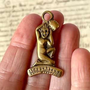 May include: A brass-coloured metal pendant featuring a sculpted figure of a seated character holding a lantern above its head. The base of the pendant is a curved banner with the embossed text JACK O LANTERN. The piece includes a circular loop at the top.
