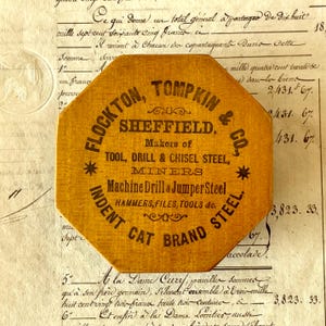 May include: An octagonal mustard-yellow wooden plaque featuring black printed text that reads: FLOCKTON, TOMPKIN & CO., SHEFFIELD. Makers of TOOL, DRILL & CHISEL STEEL, MINERS Machine Drill & Jumper Steel, HAMMERS, FILES, TOOLS &c. INDENT CAT BRAND STEEL.