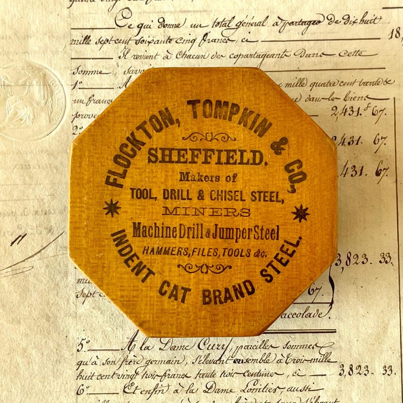 May include: An octagonal mustard-yellow wooden plaque featuring black printed text that reads: FLOCKTON, TOMPKIN & CO., SHEFFIELD. Makers of TOOL, DRILL & CHISEL STEEL, MINERS Machine Drill & Jumper Steel, HAMMERS, FILES, TOOLS &c. INDENT CAT BRAND STEEL.