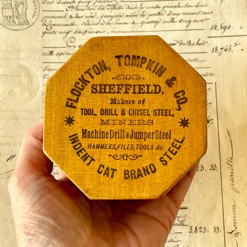 May include: An octagonal wooden box with a yellow finish and black printed text that reads FLOCKTON, TOMPKIN & CO., SHEFFIELD. Makers of TOOL, DRILL & CHISEL STEEL, MINERS, Machine Drill & Jumper Steel, HAMMERS, FILES, TOOLS & CO. INDENT CAT BRAND STEEL.
