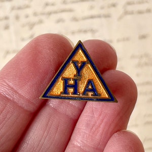 May include: A triangular pin with a gold-colored center and blue border. The pin features the letters "YHA" in blue. The pin is held in a person's hand.