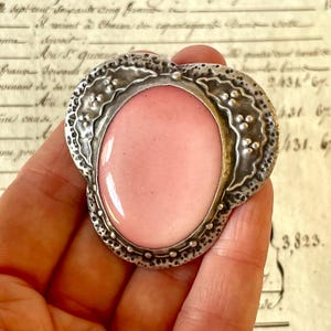 Puede incluir: Broche vintage plateado con una gran piedra ovalada de color rosa pálido. El broche tiene un borde adornado y texturizado con un diseño floral. El broche se sostiene en una mano sobre un fondo de papel envejecido.