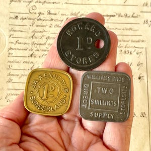 May include: Three vintage metal trade tokens. One round dark metal token reads HOWARD STORES 1P. One square brass-coloured token reads J.A. KENNEDY LTD SUNDERLAND 1P. One square dark metal token reads WILLIAMS BROS DIRECT SUPPLY STORES LTD TWO SHILLINGS.