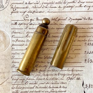 May include: A cylindrical brass case with a rounded top knob and a separate matching cap. The metal has a weathered, golden-brown patina with visible surface texture. The object is composed of two distinct pieces, a main body and a removable lid.