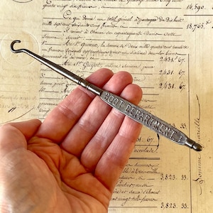 May include: A vintage metal boot hook featuring a long, slender handle with the words BOOT DEPARTMENT embossed in capital letters. The tool has a curved hook at one end and a rounded tip at the other, finished in a dark, weathered metallic tone.