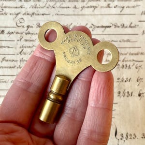 May include: A vintage brass clock key with a distinctive spade symbol and the text "MADE IN POPULAR ENGLAND PROGRESS." The key has a cylindrical stem and two circular openings. The key is a warm gold color.