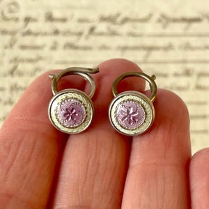 Op de afbeelding: Twee antieke manchetknopen in zilverkleur met een bloemmotief in roze en wit email. De manchetknopen hebben een cirkelvorm met een decoratieve rand en een gebogen staaf. De achtergrond is vintage papier.