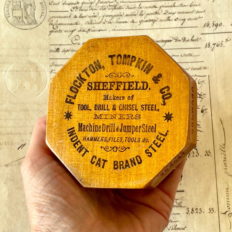 May include: An octagonal wooden box with a yellow finish and black printed text that reads Flockton, Tompkin & Co., Sheffield, Makers of Tool, Drill & Chisel Steel, Miners, Machine Drill & Jumper Steel, Hammers, Files, Tools & Co., Indent Cat Brand Steel.