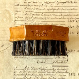 May include: A wooden brush with a rectangular handle featuring the embossed text WANKLYN'S PATENT and REG D NO 383041. The brush has dense, stiff, dark brown and black bristles arranged in rows, providing a functional tool for cleaning or grooming.