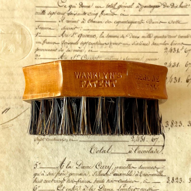 May include: A wooden brush with a rectangular handle featuring the embossed text WANKLYN'S PATENT and REG D NO 383041. The brush has dense, stiff, dark brown and black bristles arranged in rows, providing a functional tool for cleaning or grooming.