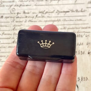 May include: A small, rectangular, black hinged box with a glossy finish. The lid features a centred, gold-coloured graphic of a five-pointed crown. The box is compact and designed for storage, with a smooth surface and clean, rounded edges.