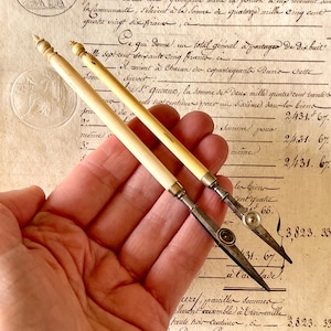 May include: Two antique writing instruments featuring long, cream-coloured cylindrical handles with pointed finials. Each tool has a metallic ferrule connecting the handle to a sharp, pointed steel nib, with one nib featuring a small circular decorative element.