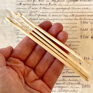 May include: A set of four off-white, smooth, cylindrical crochet hooks with tapered ends. Each hook features a distinct notch near the tip for yarn manipulation. The tools are arranged in a parallel formation, showcasing a uniform, light-colored finish.