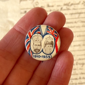 Puede incluir: Un botón redondo con fondo blanco y un diseño de Union Jack rojo, blanco y azul. El botón presenta una imagen en blanco y negro del rey Jorge V y la reina María, con el texto "1910-1935" debajo.
