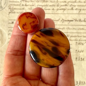 May include: Two circular buttons featuring a glossy, marbled tortoiseshell pattern in shades of amber, brown, and black. The buttons are smooth, round, and flat, displaying a distinct swirling color variation across their polished surfaces.