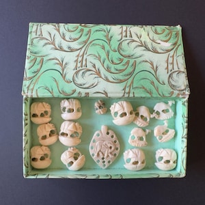 May include: A light green cardboard box with a dark green fern-like pattern containing twelve small, off-white carved elephant figurines and one larger, flat, leaf-shaped pendant featuring a central elephant carving with multiple circular cutouts.