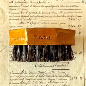 May include: A rectangular wooden brush with dark, stiff bristles. The side of the light brown wooden handle features the engraved text OBLWARA and T G.O.L. The brush has a smooth, polished finish and a flat top surface for a firm grip during use.